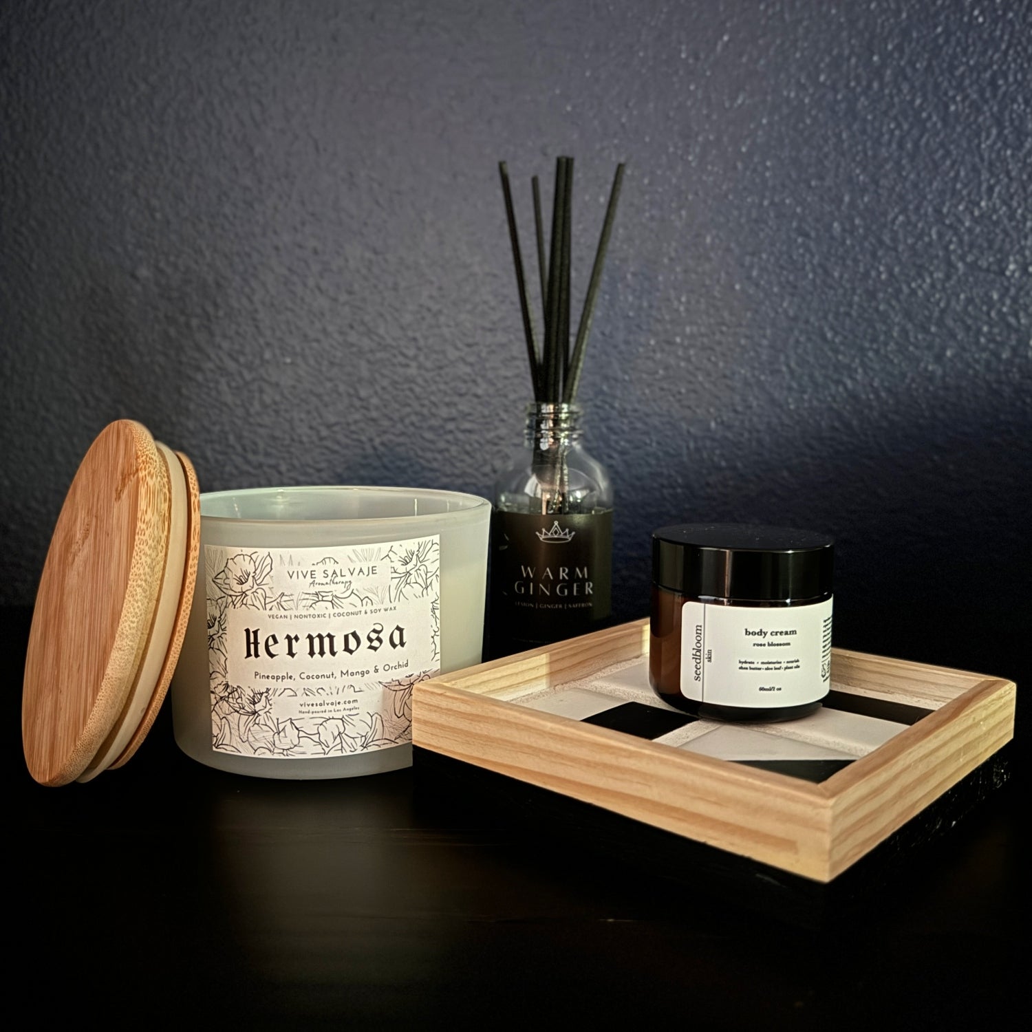 Collection of bedside aromatherapy items: three wick candle, warm ginger diffuser, rose blossom lotion, and a tile tray on a dark table with a blue background
