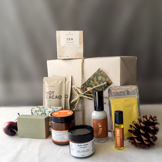 Collection of wellness products in the New Mom Self-Care Gift Box, including hot cacao, a turmeric face mask, and various other skincare products in front of a wrapped gift box. 