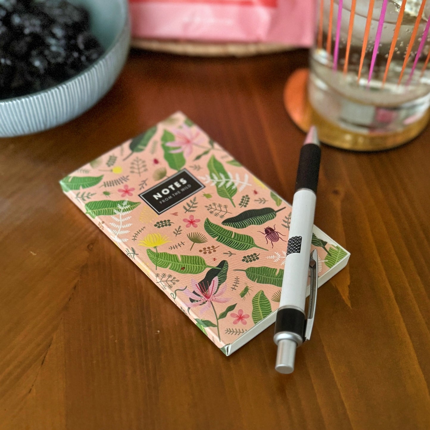 Small notebook with pink and green motif on table with pen and a glass