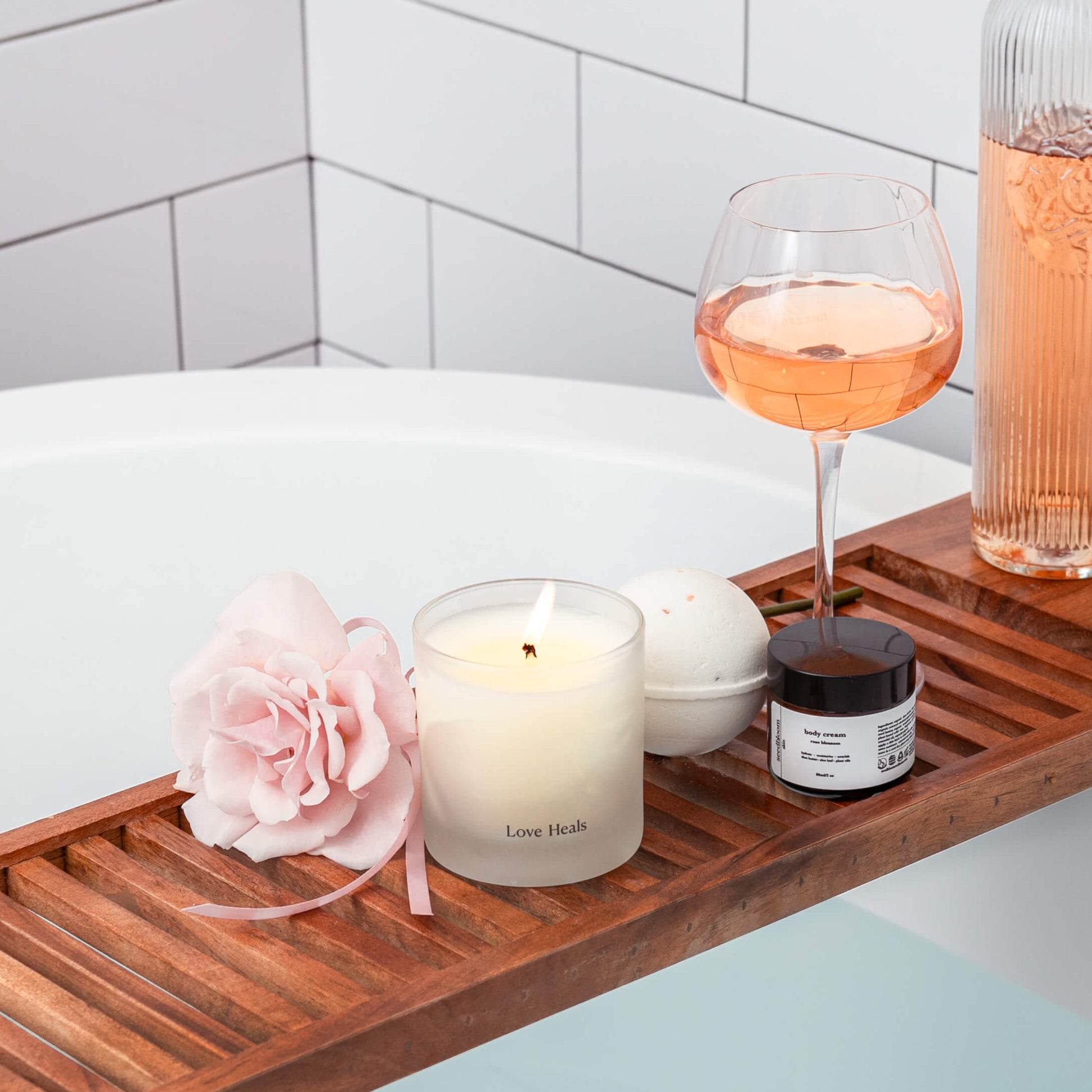 A Valentine gift that gives back on a wooden bath tray with a candle, flower, glass of rose, and jar of rose lotion.