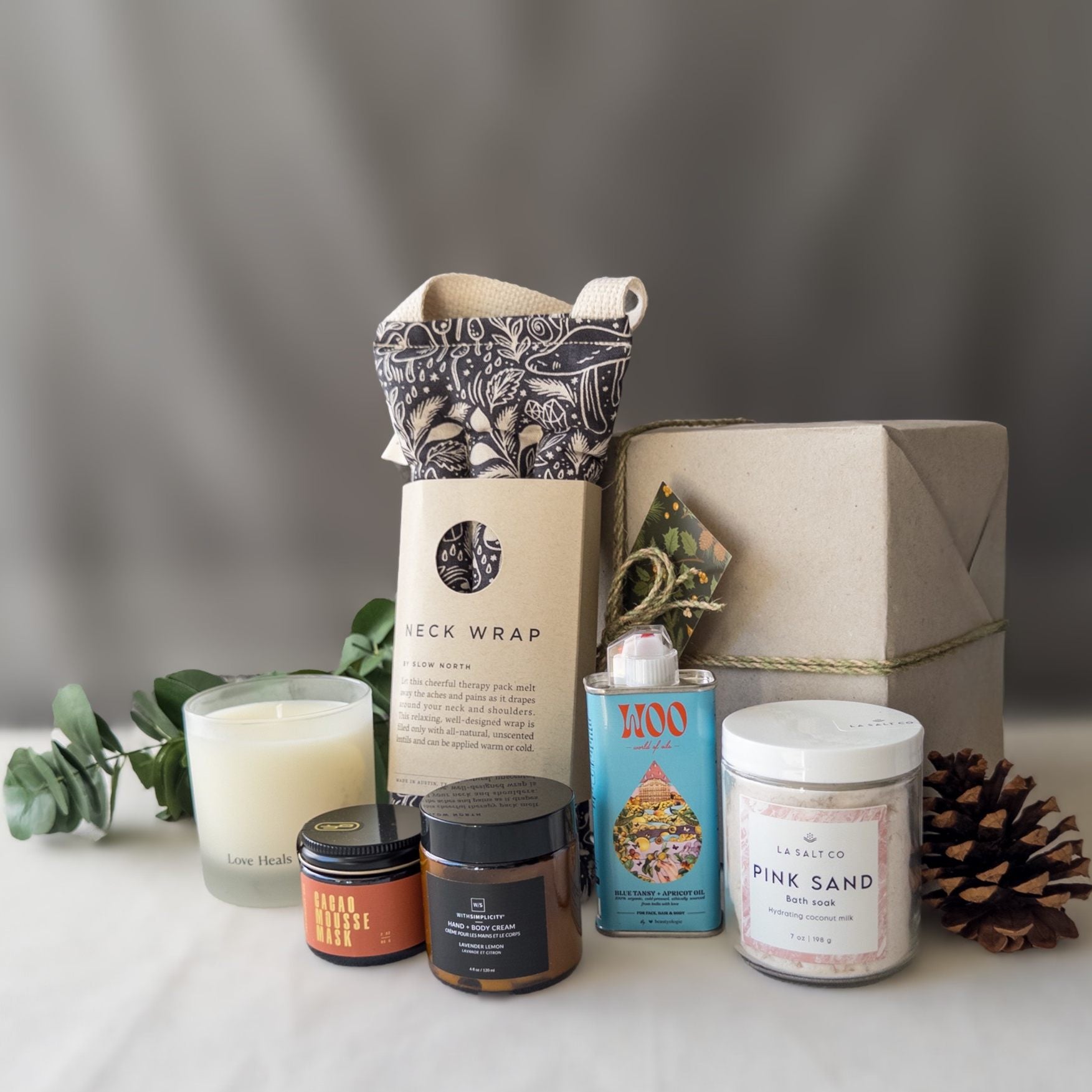 Contents of the Rest & Relaxation Gift Box, including weighted neck wrap, candle, skincare, and Pink Sand bath soak in front of a wrapped gift box with pinecone.
