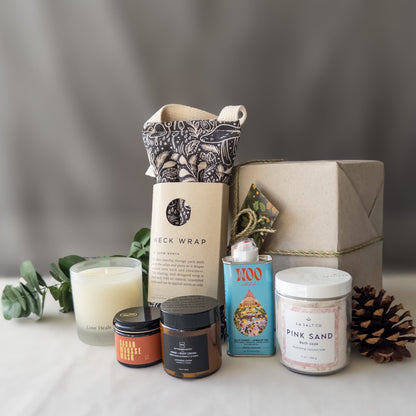 Contents of the Rest & Relaxation Gift Box, including weighted neck wrap, candle, skincare, and Pink Sand bath soak in front of a wrapped gift box with pinecone.