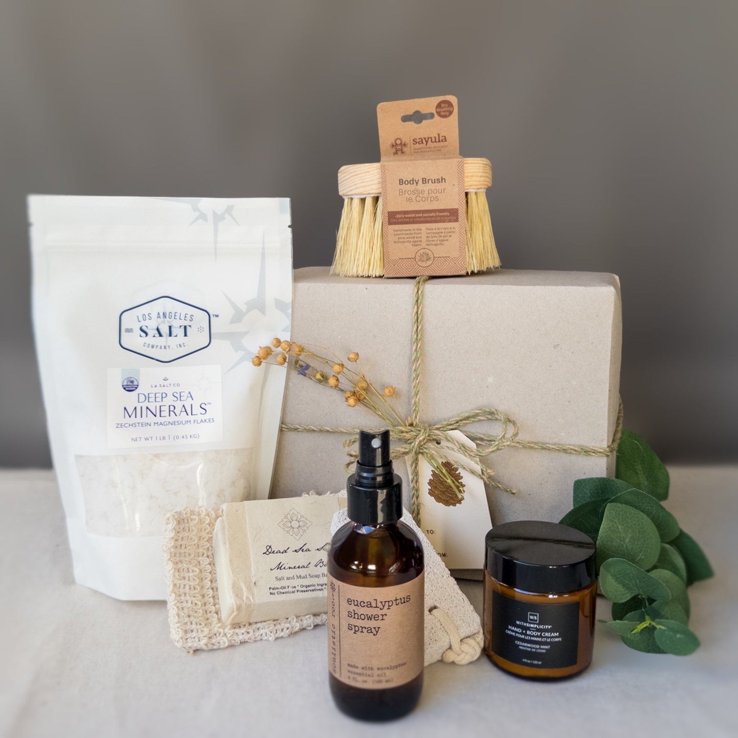 Contents from the scrub box including sea salts, body brush, shower spray, and lotion in front of a wrapped gift box with fresh eucalyptus.