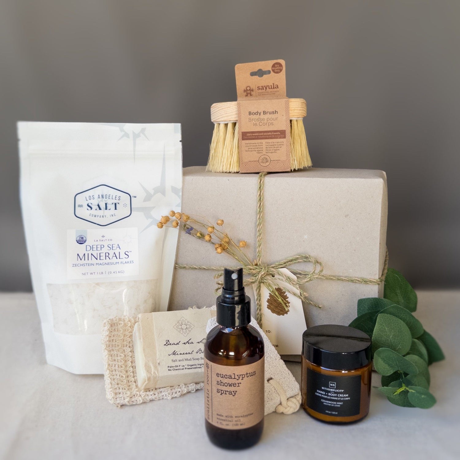 Contents from the scrub box including sea salts, body brush, shower spray, and lotion in front of a wrapped gift box with fresh eucalyptus.