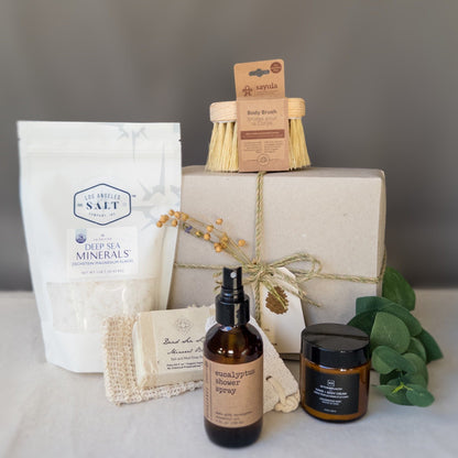 Contents from the scrub box including sea salts, body brush, shower spray, and lotion in front of a wrapped gift box with fresh eucalyptus.