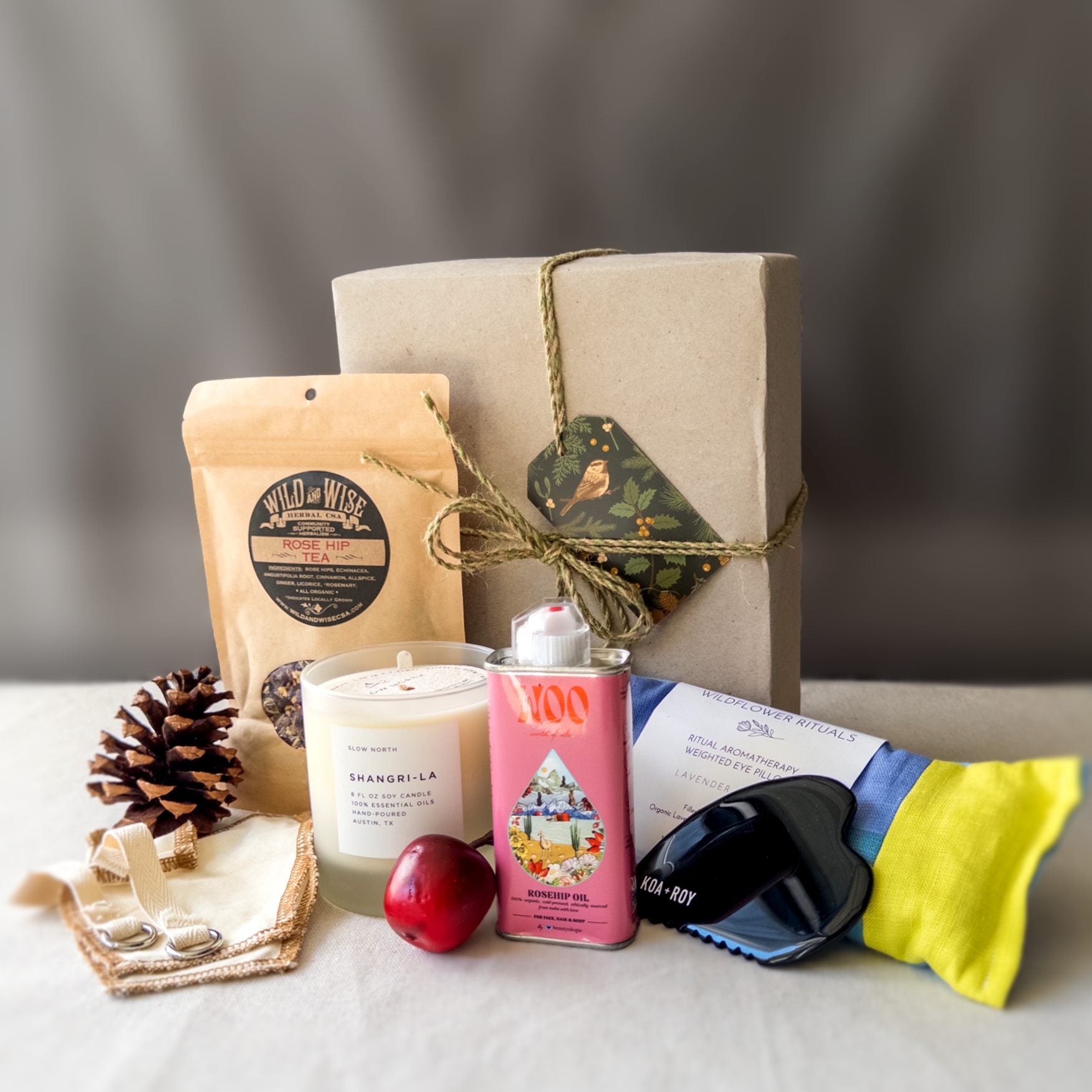 Stress relief care package contents with candle, eye pillow, tea, gua sha and body oil in front of a wrapped gift box.