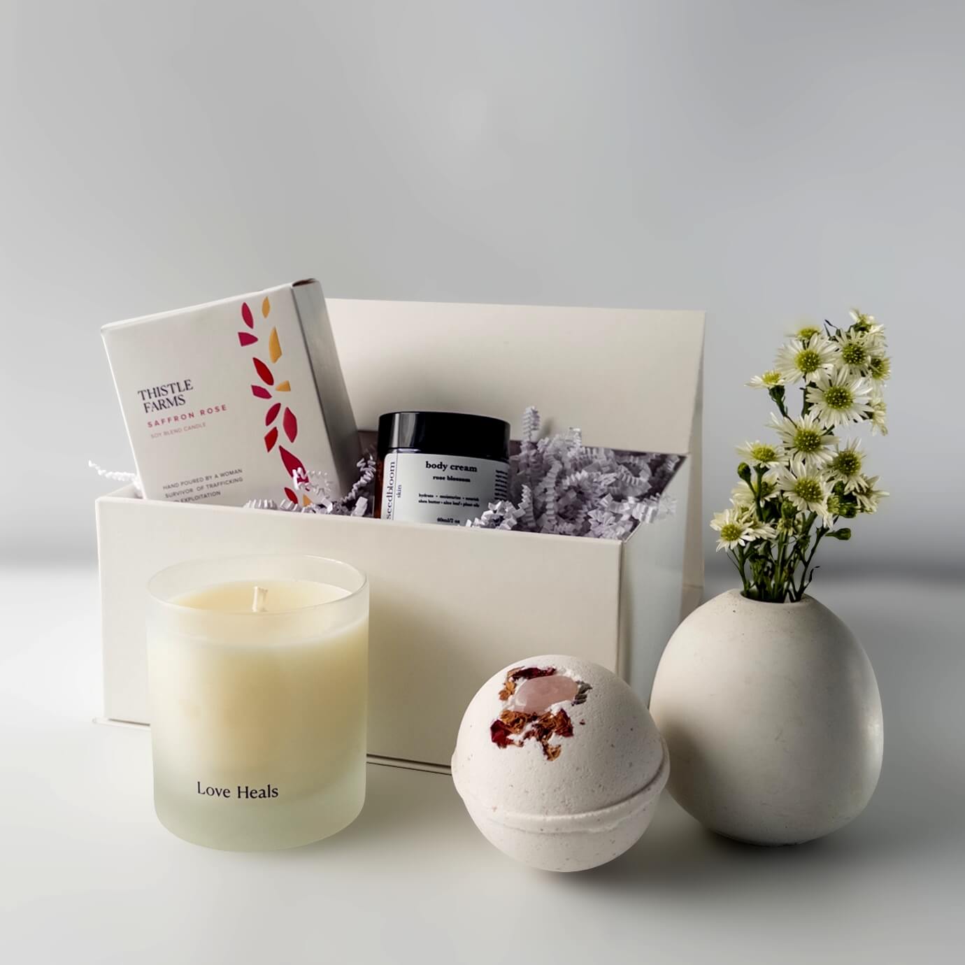 A Valentine's Day gift box with candle, rose lotion and a rose bath bomb in a white box next to flowers