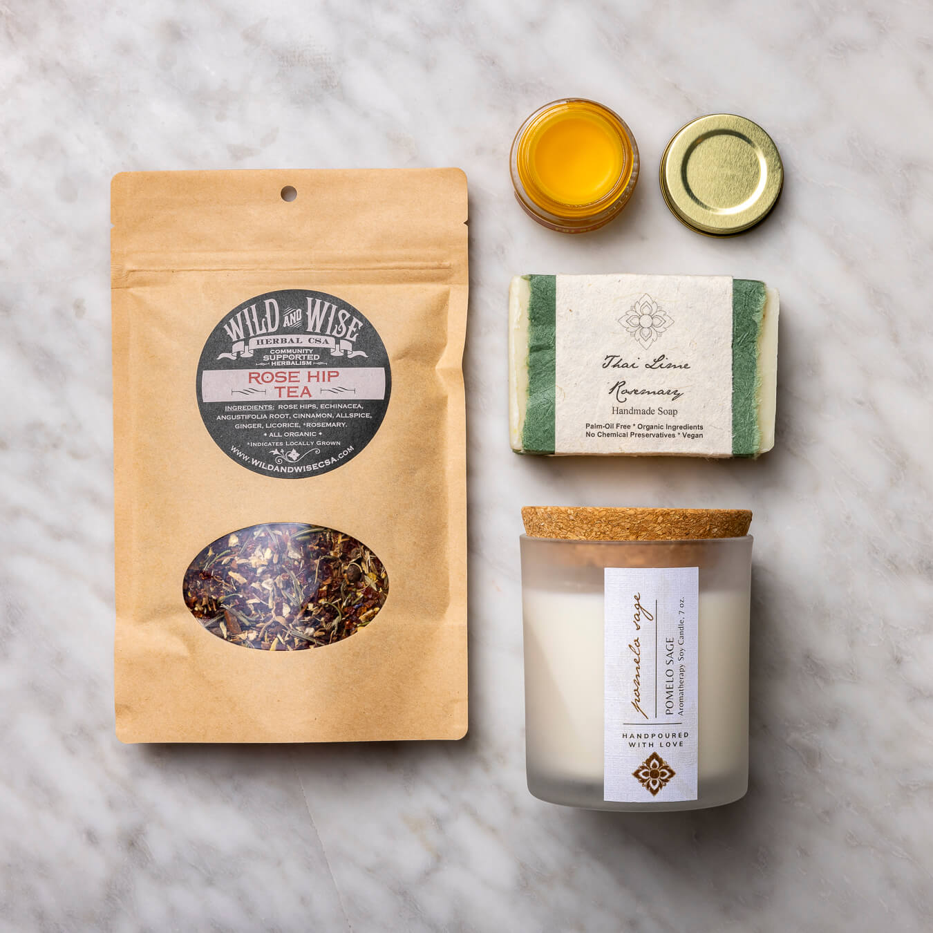 Pomelo Sage Candle, Pot of Gold Face Balm, Thai Lime Rosemary Soap, and Rosehip tea on white background.