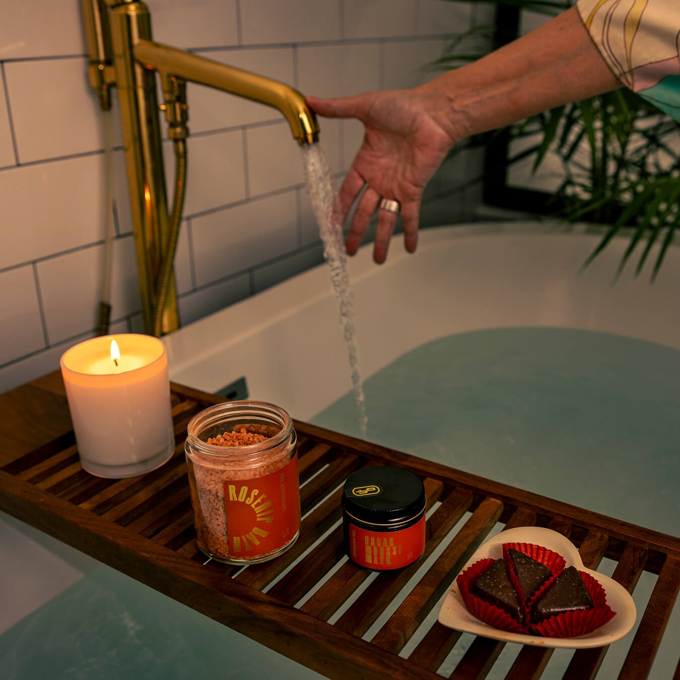 A serene setting in a bathroom with a candle, bath salts, a jar of cacao mousse mask, and three chocolate truffles over a large bath tub that is filling with water