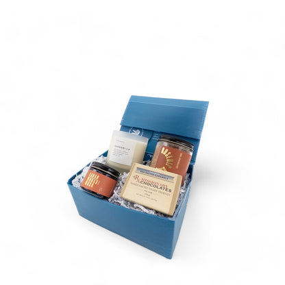 The items in Blissful Bath Box, including Cacao Mousse Mask, Rosehip Bath, Shangri-La candle, and vanilla salted caramels, in a box with white crinkle paper