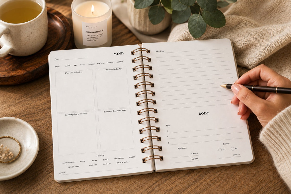 An open spiral-bound journal with daily prompts on lined pages lies on a wooden table, with a hand holding a pen writing in the body section. A mug of tea, a lit candle, a small dish, and a knit sweater are visible around it in a cozy indoor setting.