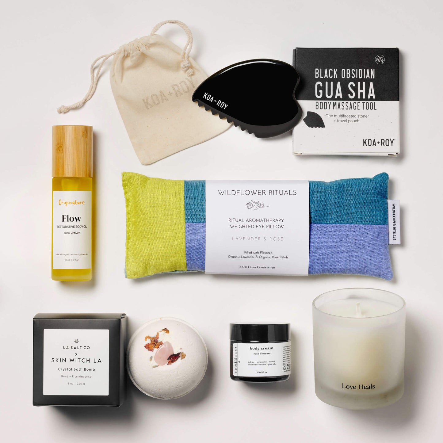 Collection of wellness products from the Goddess Luxury Spa Box, including a weighted eye pillow, candle, and skincare items on a white tile surface.
