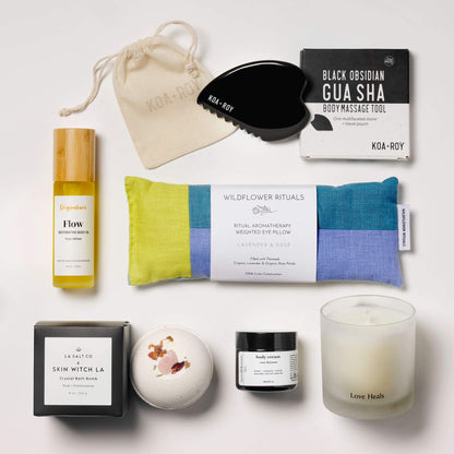 Collection of wellness products from the Goddess Luxury Spa Box, including a weighted eye pillow, candle, and skincare items on a white tile surface.