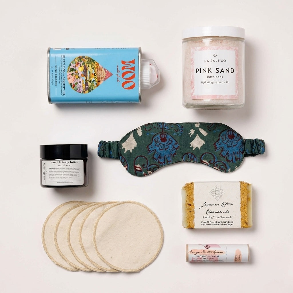 A curated postpartum gift box for new moms arranged on a  white background, with a tin of blue tansy face oil, a jar of Pink Sand bath soak, a green sleep mask, a small black jar of body lotion, a stack of reusable washcloths, a bar of Chamomile soap, and lip balm.