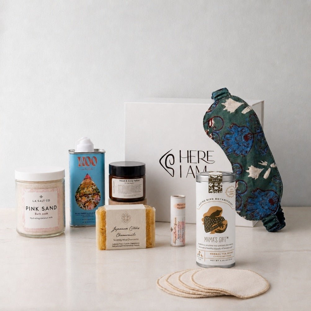 A curated wellness gift set arranged on a light surface against a pale wall. The set includes a jar of Pink Sand bath soak, a blue tin of Blue Tansy oil, a small dark jar of rose blossom lotion, a bar of handmade soap, a small tube of lip balm, a can of herbal tea for nursing mothers, with a white gift box with the Here I Am logo, and a patterned green sleep mask.