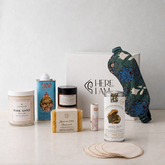 A curated wellness gift set arranged on a light surface against a pale wall. The set includes a jar of Pink Sand bath soak, a blue tin of Blue Tansy oil, a small dark jar of rose blossom lotion, a bar of handmade soap, a small tube of lip balm, a can of herbal tea for nursing mothers, with a white gift box with the Here I Am logo, and a patterned green sleep mask.