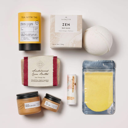 Collection of wellness products in a box for new moms, including sandalwood soap, a turmeric face mask, and various other items on a light background.
