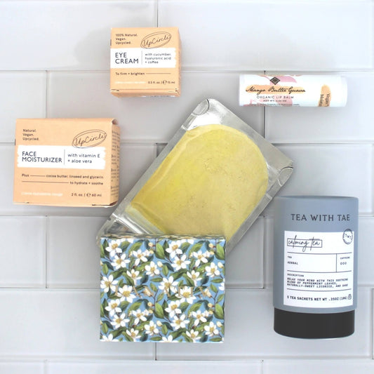 Collection of wellness products in the New Mom Self-Care Gift Box, including sandalwood soap, a turmeric face mask, and various other items on a light background.