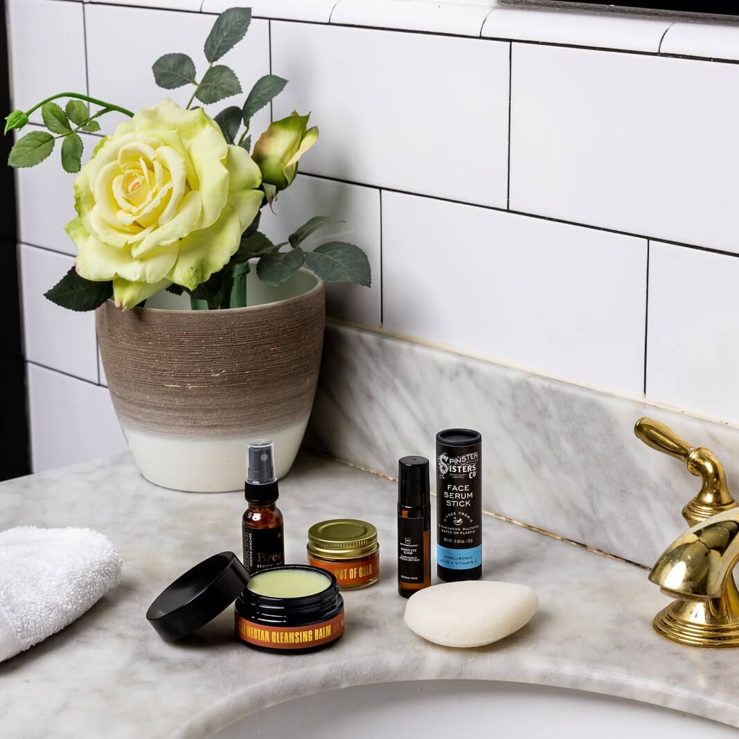 Pot of Gold Regenerative Face Balm, Breeze Toner, Konjac sponge, Goji Nectar Cleanser, Face Serum stick, and Eye Elixir on bathroom counter with rose