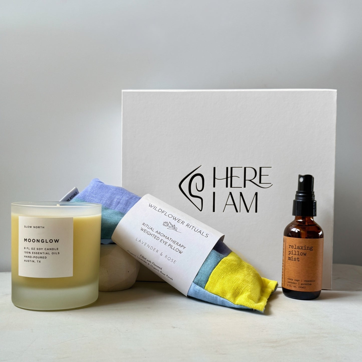 A curated wellness gift set featuring a frosted glass candle, a relaxing eye pillow, and a small amber bottle of pillow spray, arranged around a white gift box on a light surface against a soft neutral background.