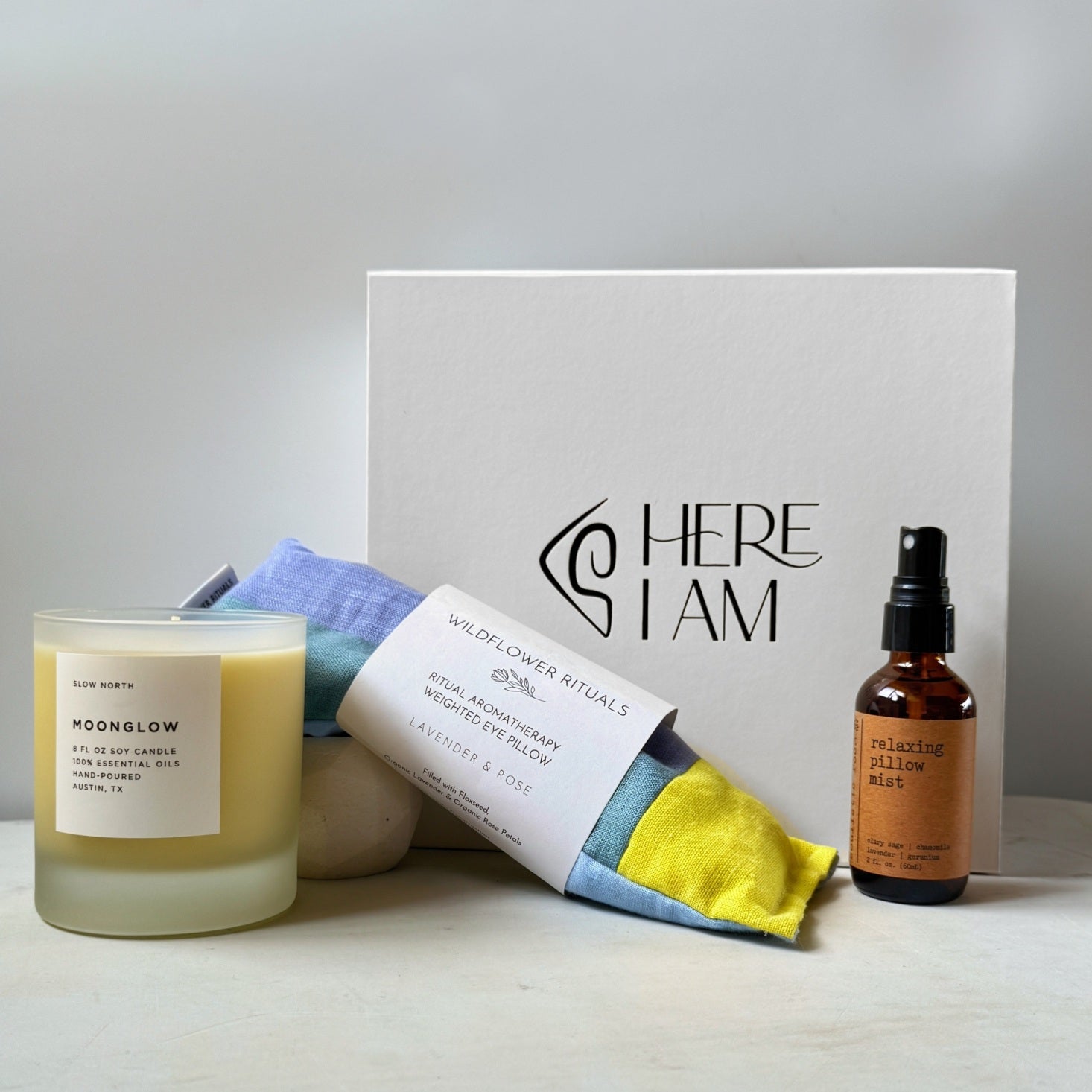 A curated wellness gift set featuring a frosted glass candle, a relaxing eye pillow, and a small amber bottle of pillow spray, arranged around a white gift box on a light surface against a soft neutral background.