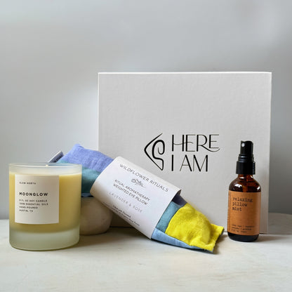 A curated wellness gift set featuring a frosted glass candle, a relaxing eye pillow, and a small amber bottle of pillow spray, arranged around a white gift box on a light surface against a soft neutral background.