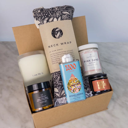 Contents of the Rest & Relaxation Gift Box in a box on a counter, including weighted neck wrap, candle, lotion, and Pink Sand bath soak