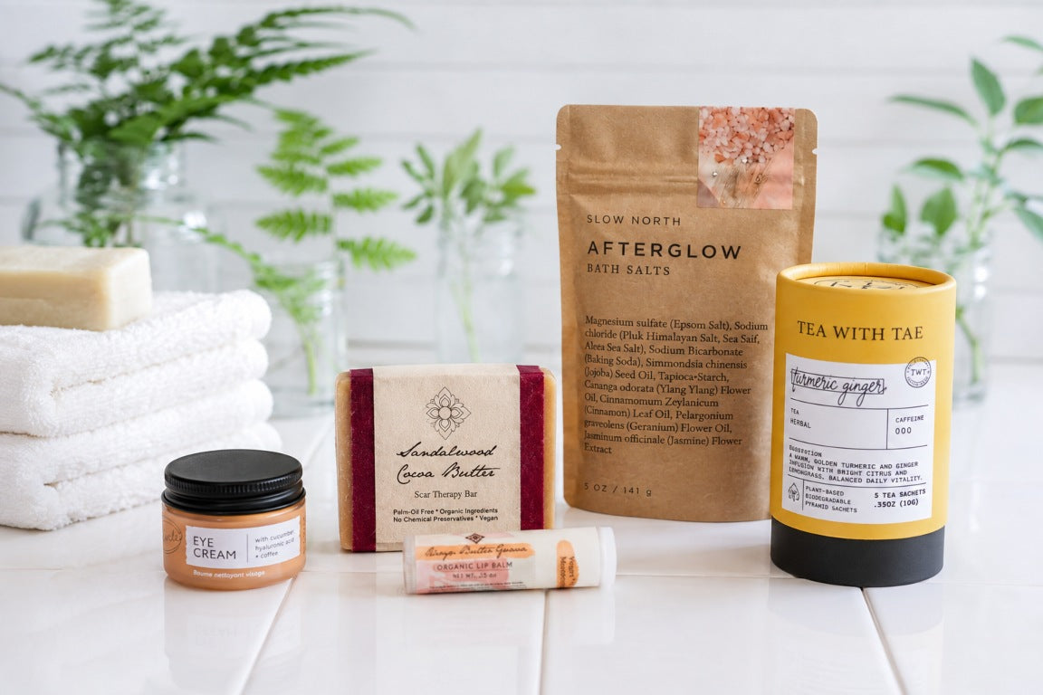 A curated collection of self-care and beauty products from the Skin Renewal Self-Care Gift Box arranged on a white tile surface with folded white towels and green plants in the background. The products include a jar of eye cream, a bar of sandalwood soap, a tube of lip balm, a brown pouch with magnesium bath salts, and a yellow tube of Turmeric Ginger Tea.