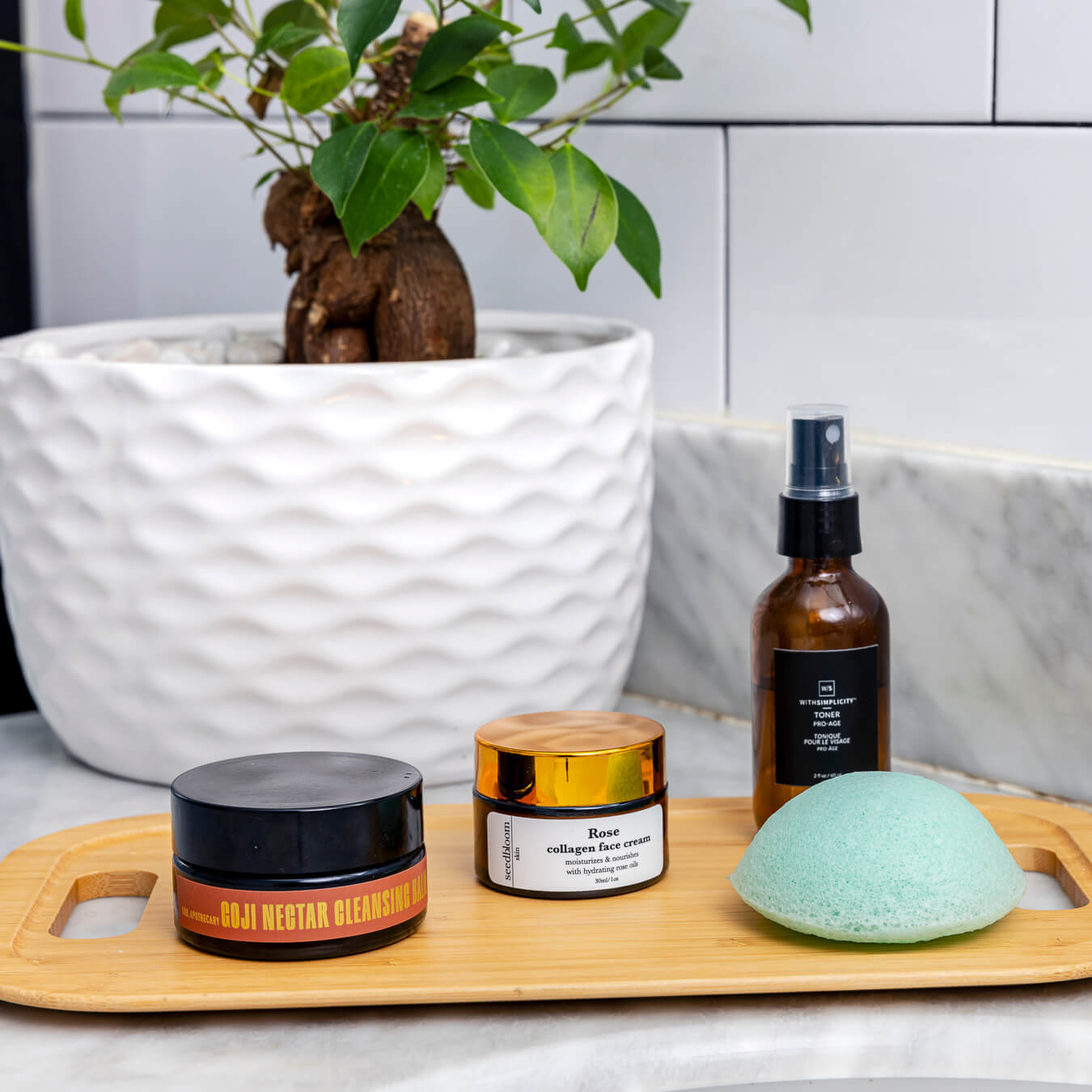 Products from the Rise and Shine box, including the Goji nectar cleansing balm, Pro-Age toner, Rose collagen face cream, and konjac sponge sitting on a bamboo tray in front of a leafy green plant