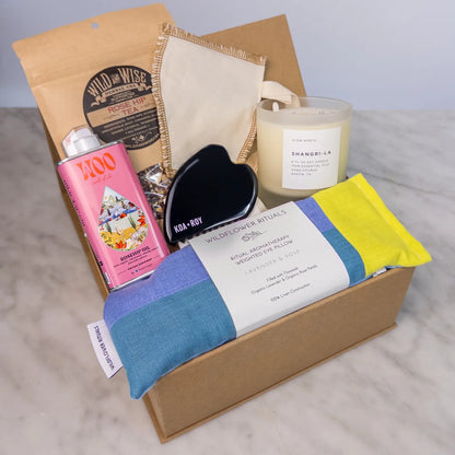 Stress relief care package in a box on a counter with candle, eye pillow, tea, gua sha and body oil. 