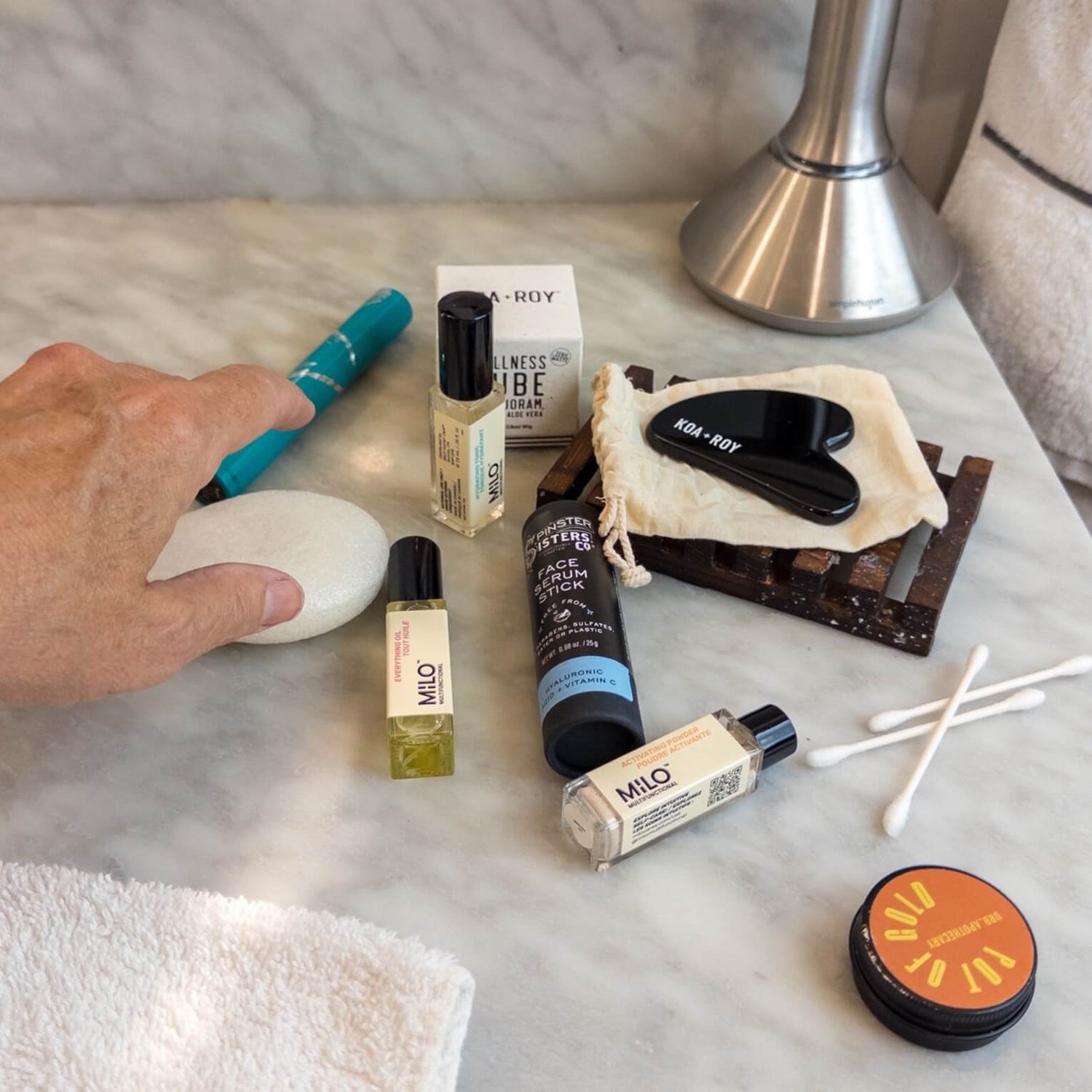 Travel sized self-care including MILO skincare, gua sha, and konjac sponge on a bathroom counter.