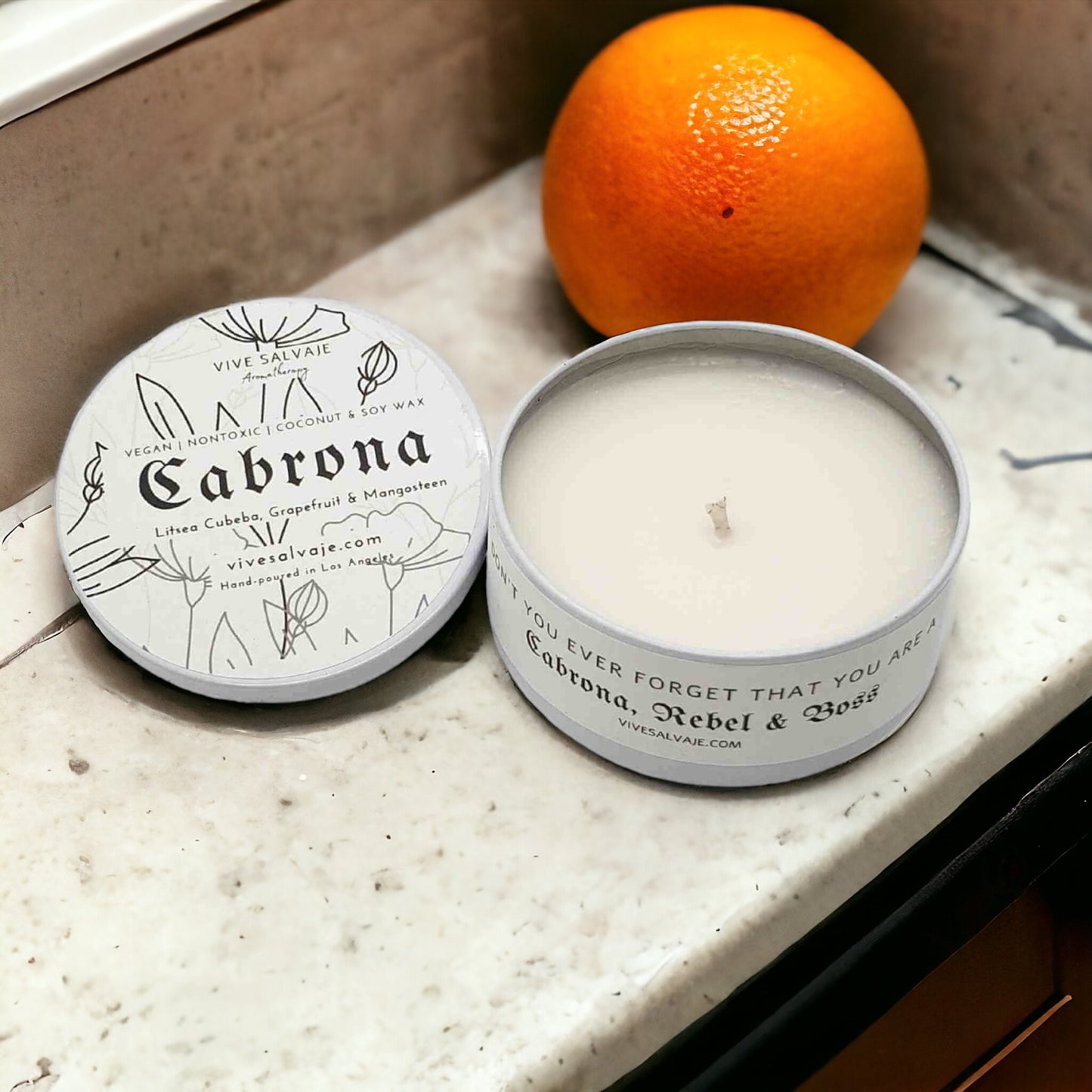 The Cabrona candle with the lid on the side next to an orange on a granite counter