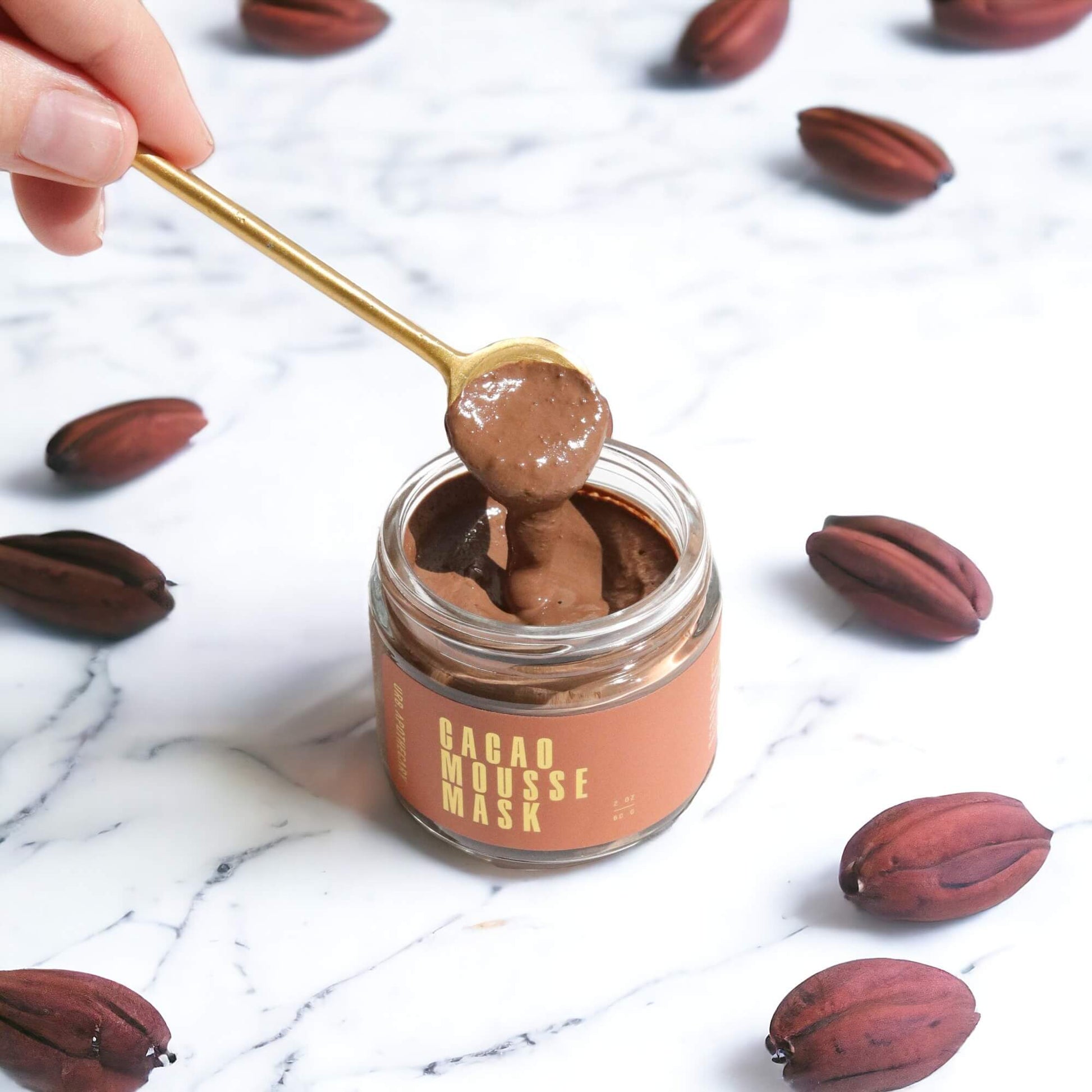 Open jar of the Cacao Mousse Mask with a gold spoonful of the mousse on a marble counter with cacao beans