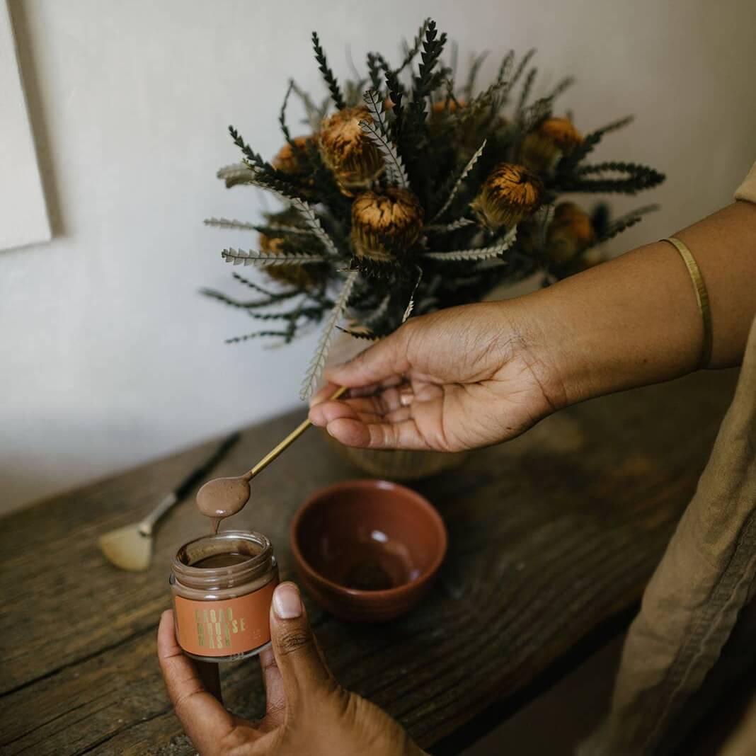 Woman with a spoonful of the Cacao Mousse Mask with a bowl and flowers on a wooden counter