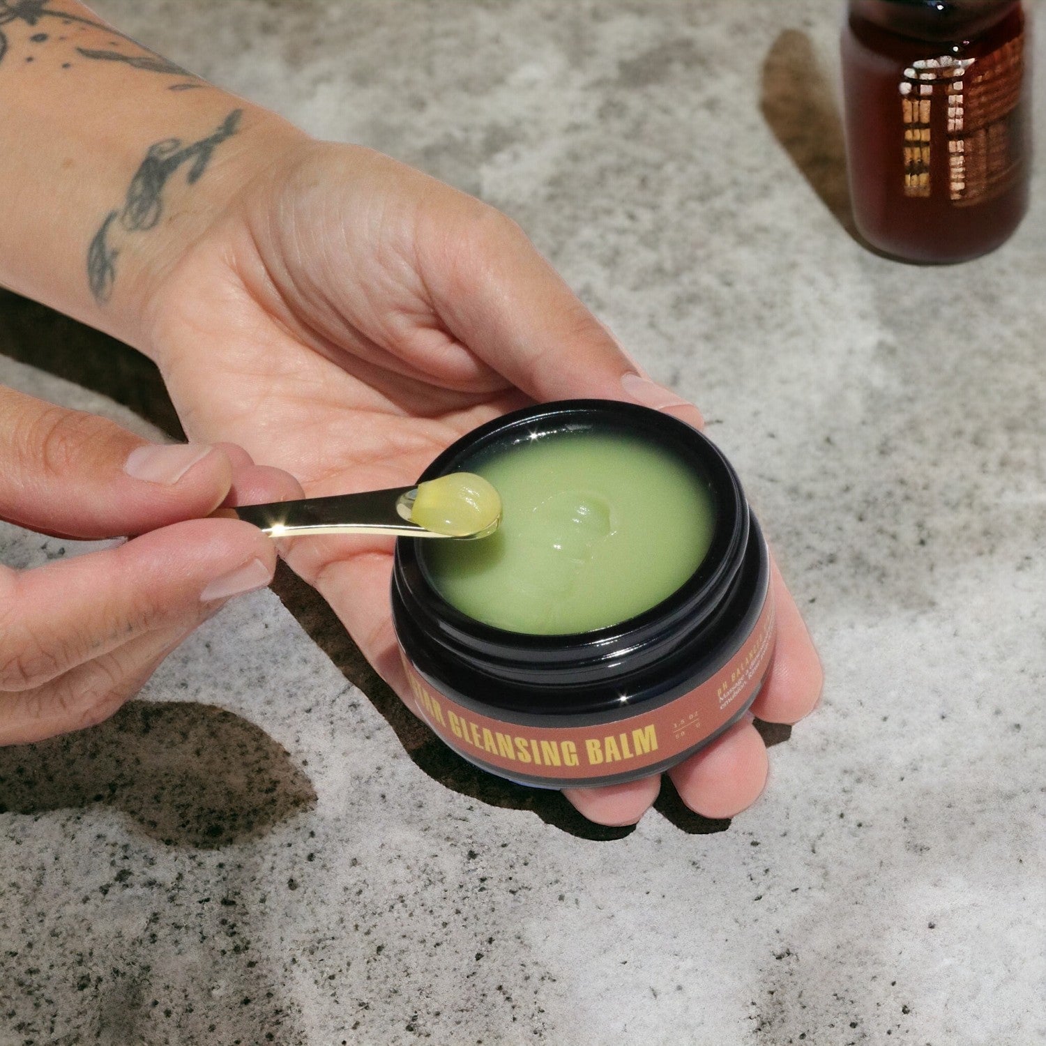 Hands holding a jar and teaspoon of the Goji Cleansing balm over a concrete countertop