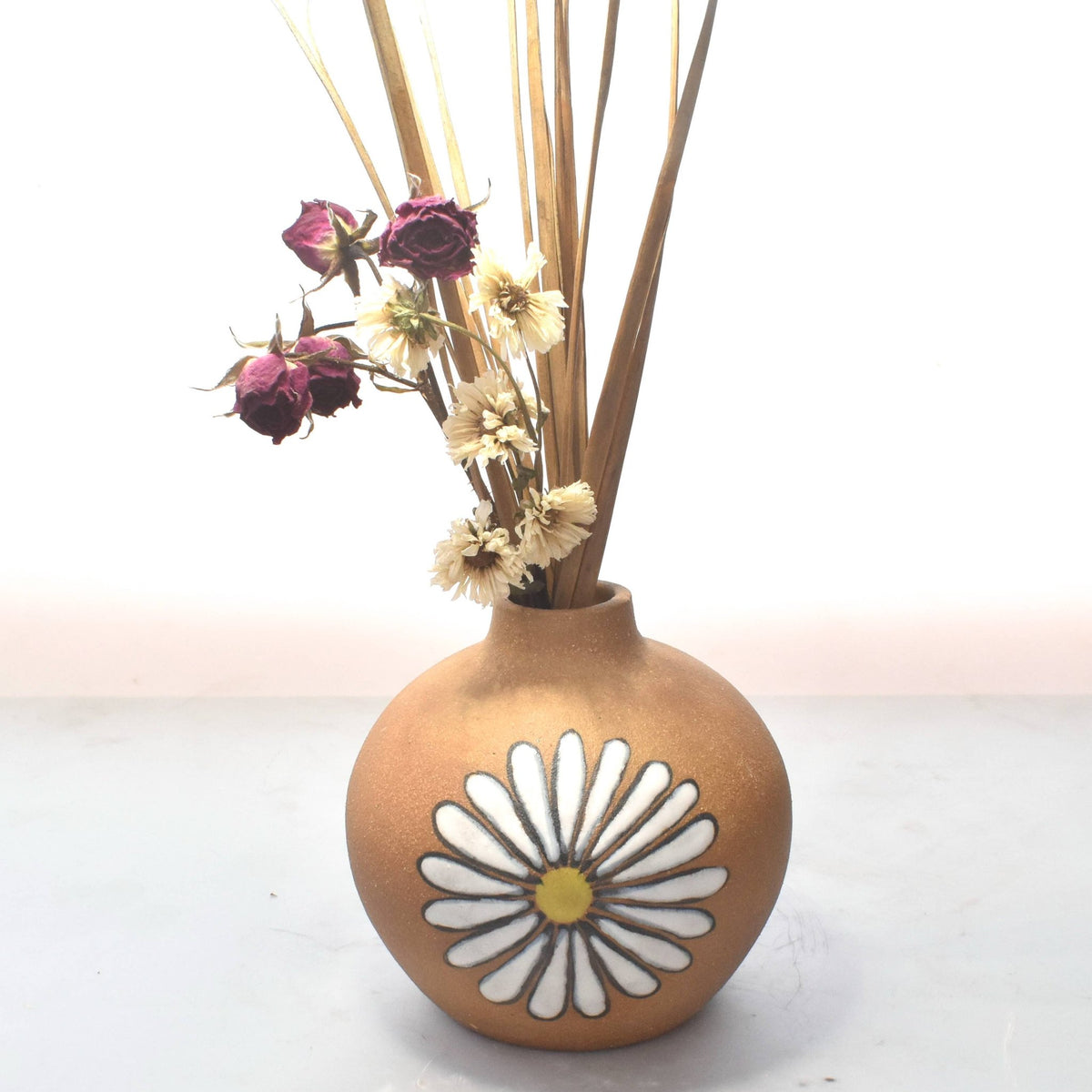 A round, matte brown ceramic vase with a white daisy design and yellow center, holding dried flowers and grass stems, set against a white background.