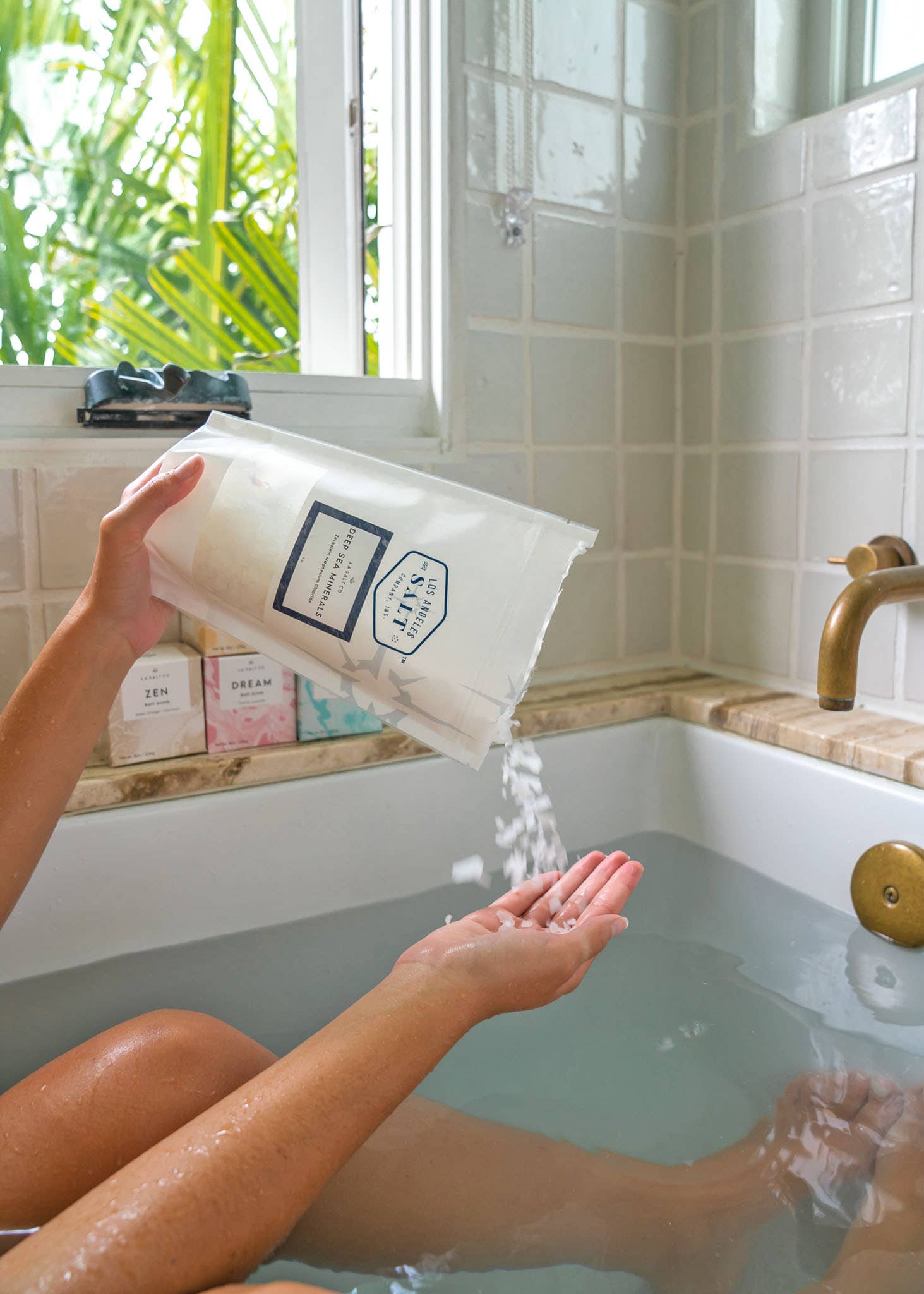 Person pouring magnesium bath flakes into a bathtub with a view of greenery outside.