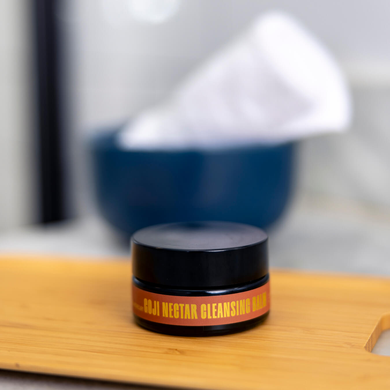 Jar of the Goji Nectar Cleansing Balm on a bamboo tray in front of a blue bowl with white towels