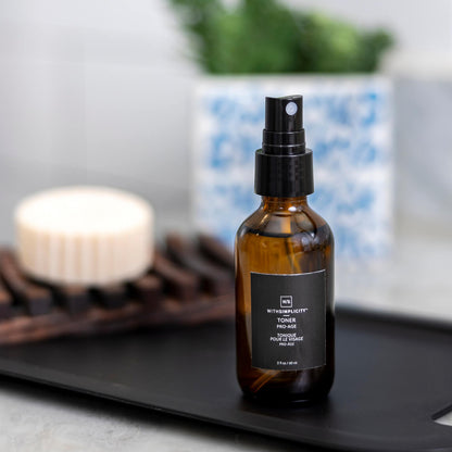 A spray bottle of the Pro-Age Toner on a sink counter with a bar of soap and green plant in the background