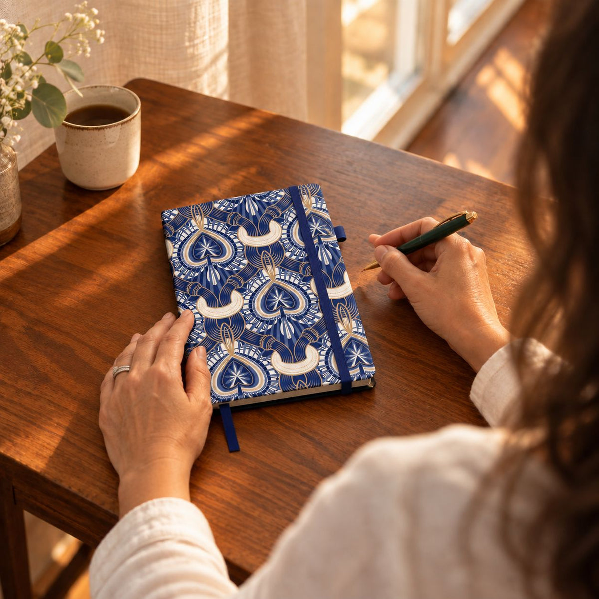 Over the shoulder of a woman, the stone paper journal sits on a small writing table near a sunny corner.