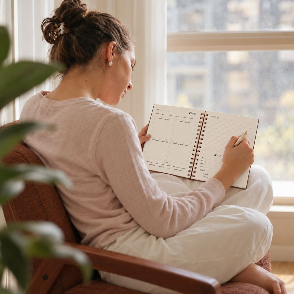 A woman sits in a brown armchair by a window, writing in the daily wellness journal with a pen. She is wearing a light pink long-sleeve top and white pants.