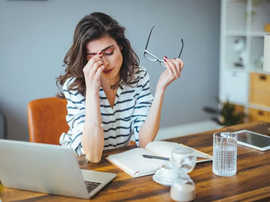 Woman sitting at a desk with a laptop, holding eyeglasses and rubbing her eyes, indicating stress.
