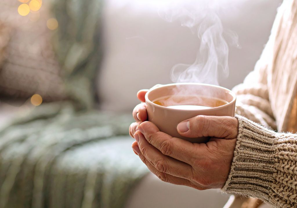 A close-up of a person's hands holding a warm ceramic mug with steam rising from it, wearing a beige knit sweater in a cozy indoor setting.