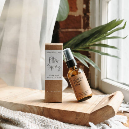 Relaxing pillow spray with lavender and chamomile and its matching kraft paper box are displayed on a wooden tray in a bright, softly lit room with a window, brick wall, and green plant leaves in the background.