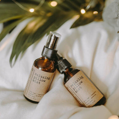 Two bottles of the lavender pillow spray with chamomile on white linens with green leaves and lights in the background.