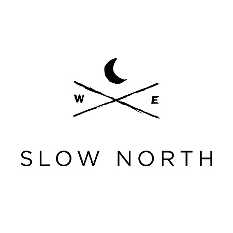 The Slow North logo on a white background featuring a crescent moon above crossed diagonal lines with the letters W and E.