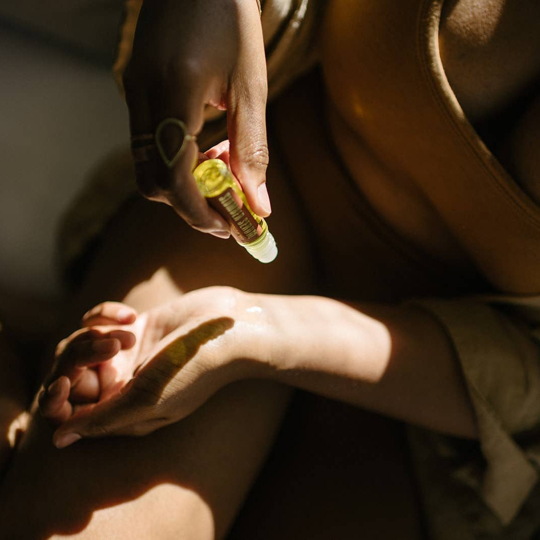 A close-up lifestyle shot of a person applying roll-on aromatherapy for sleep onto the back of their hand, with warm sunlight casting dramatic shadows.