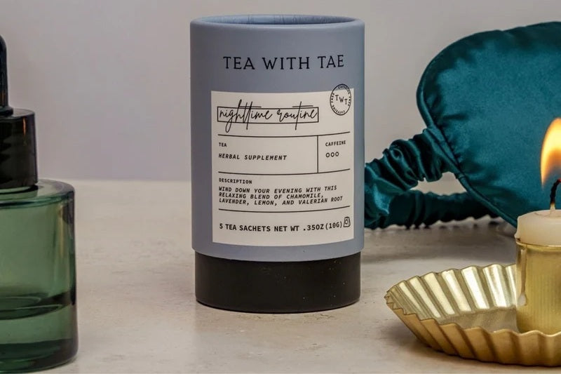 A cylindrical tube of tea on a tabletop, styled with a green glass dropper bottle on the left, a teal satin sleep mask, and a lit candle in a gold scalloped holder on the right.