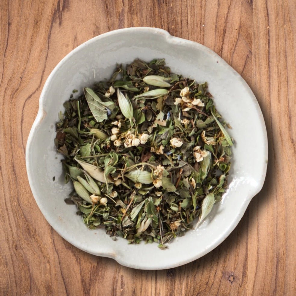 A top-down view of the Peace of Mind loose-leaf herbal tea with green leaves and small white flower buds, served in a light gray ceramic bowl on a warm brown wooden surface.