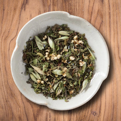 A top-down view of the Peace of Mind loose-leaf herbal tea with green leaves and small white flower buds, served in a light gray ceramic bowl on a warm brown wooden surface.
